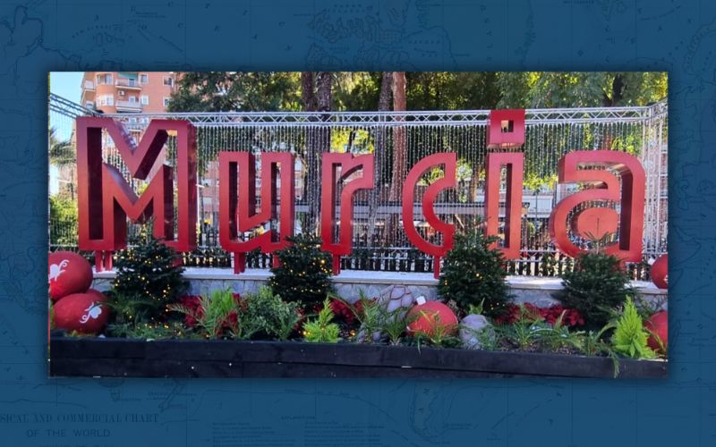The image shows a decorative installation spelling out 'Murcia' in large, bold, red letters. The letters are mounted on a metal framework and stand prominently in a public area, likely a park. The installation is adorned with green plants and small trees at its base, along with spherical red ornaments scattered around, giving it a festive look. The background suggests a sunny day with shadows cast on the ground, and there's a fence and foliage behind the sign, indicating a well-maintained urban green space.